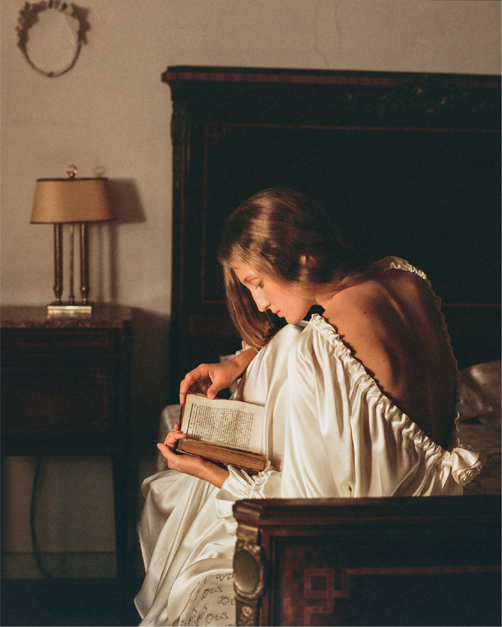 Woman in a white silk dress reading a book in a dimly lit room.