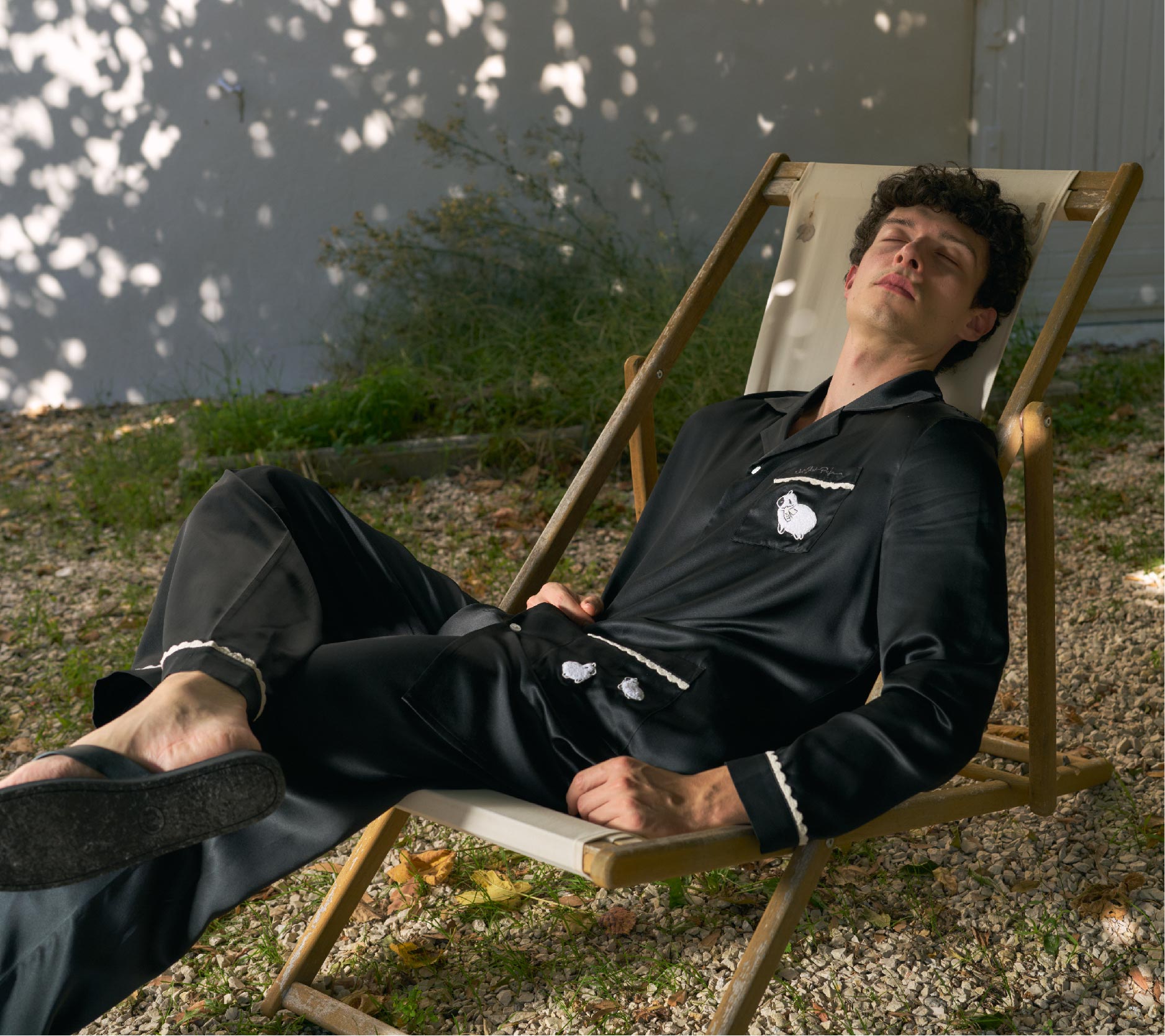 Man lounging on a wooden chair outdoors with a white wall and greenery in the background wearing Silk Pajama