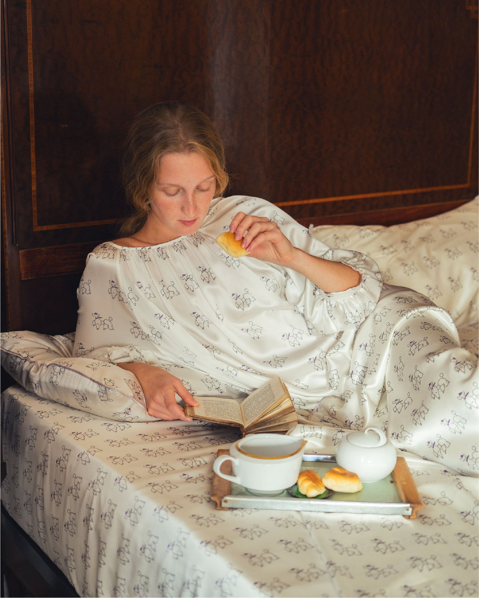 Woman wears silk pajama in bed reading a book with a tray of tea and pastries.