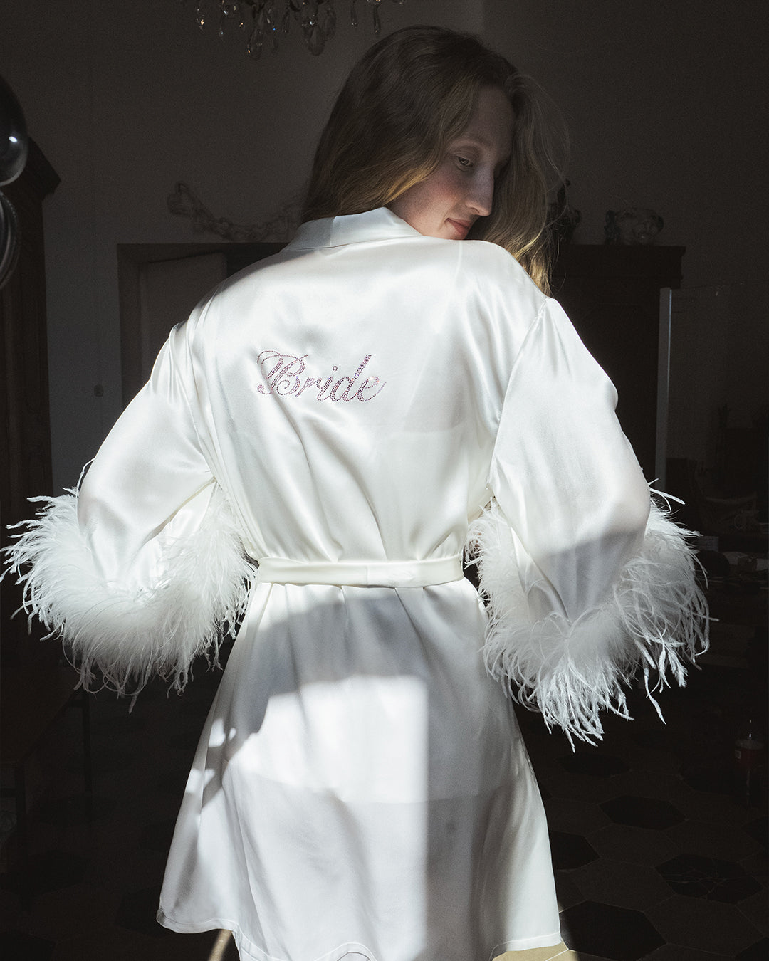 Woman wearing a white satin robe with 'Bride' embroidery and feather trim.