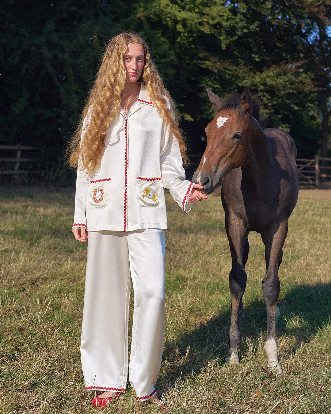 Person in a white outfit with red trim standing next to a horse in a grassy field.
