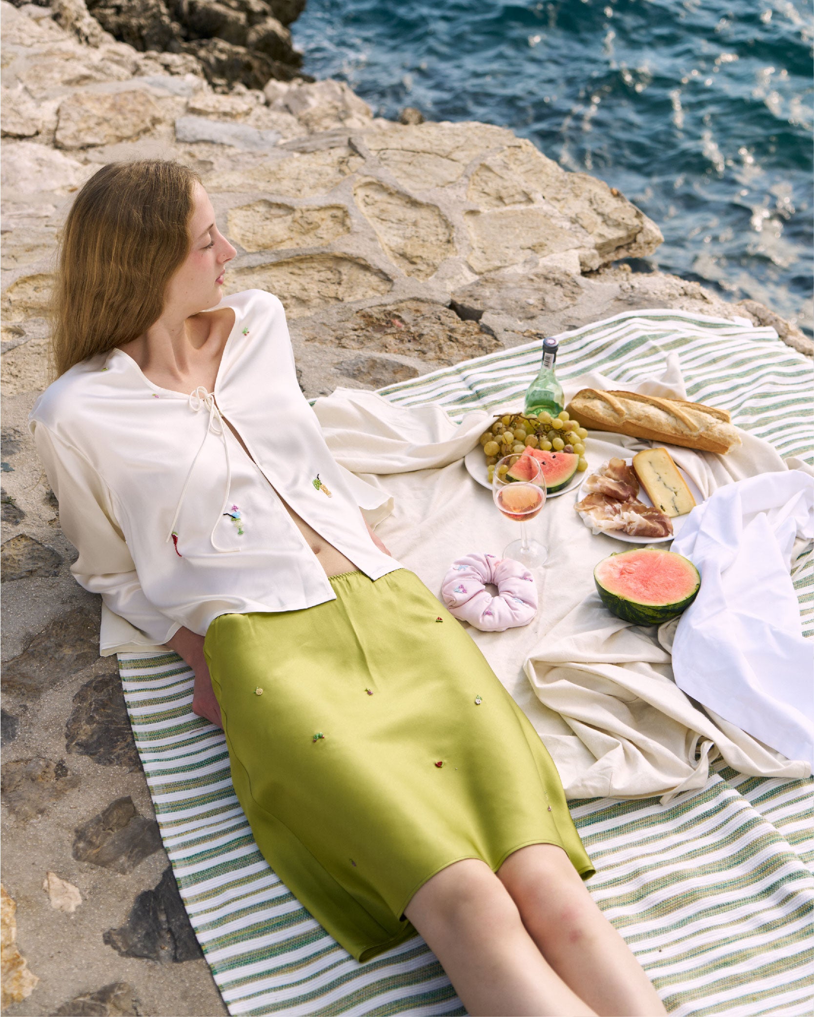Woman sitting on a striped blanket by the sea with a picnic setup.